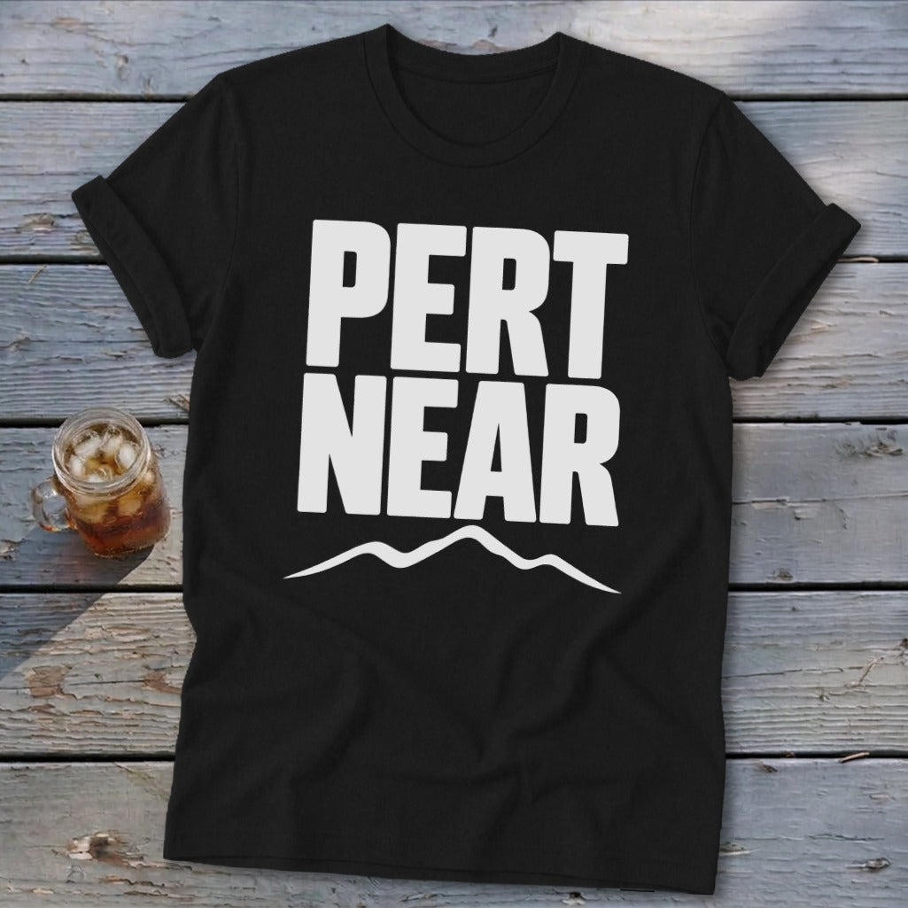 A black crewneck t-shirt featuring the white text PERT NEAR above a mountain range graphic, laid on a weathered wooden surface.
