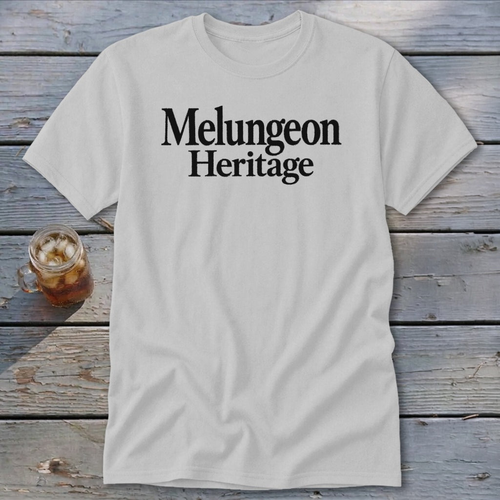 A light gray crewneck t-shirt featuring the black text Melungeon Heritage laid flat on a rustic wooden background.