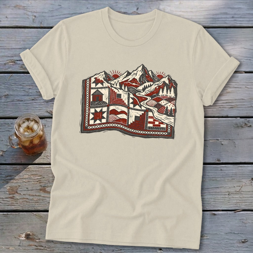A light beige t-shirt featuring a red, black, and white graphic of mountains and a quilted landscape.