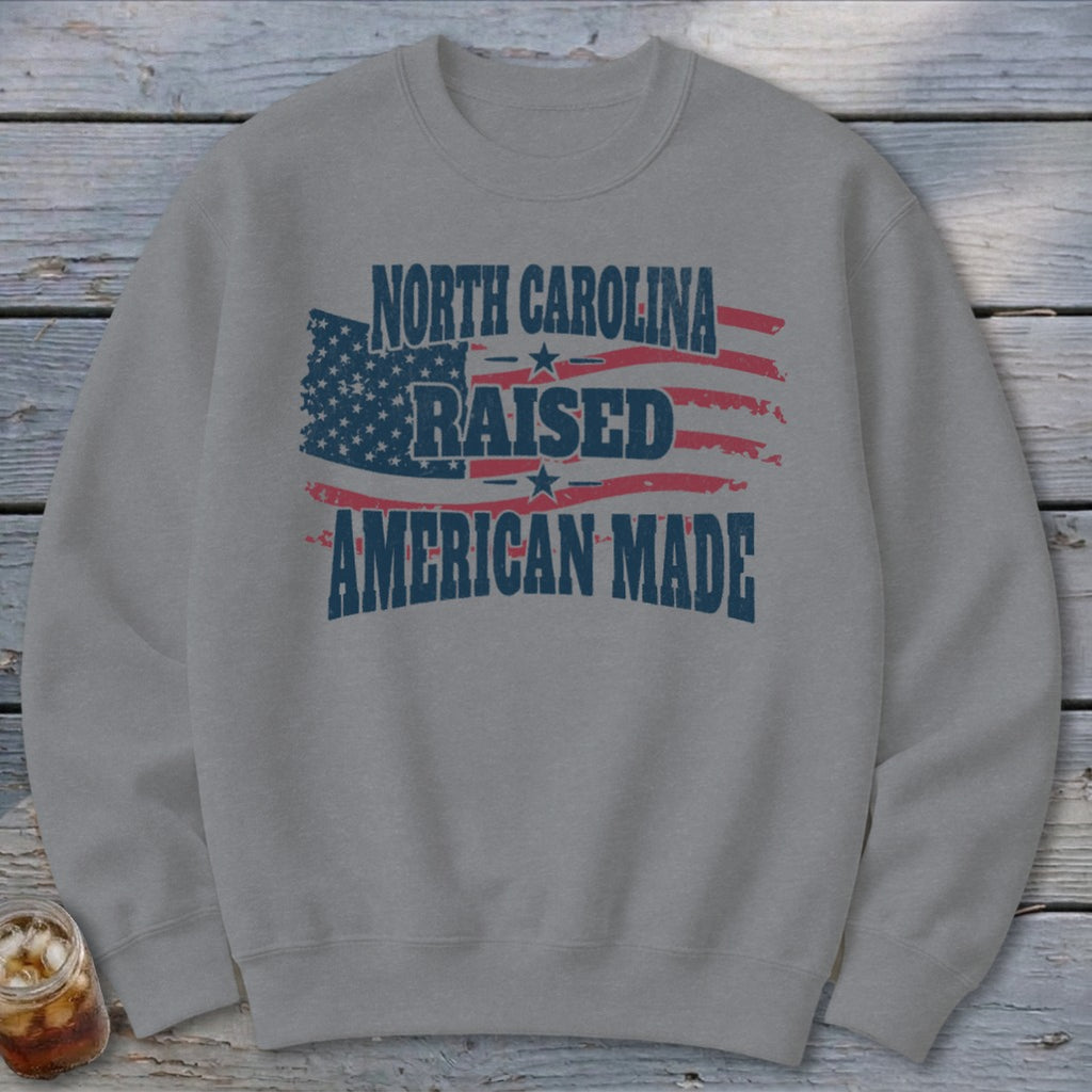 Grey crewneck sweatshirt featuring a distressed American flag graphic and the text North Carolina Raised American Made.