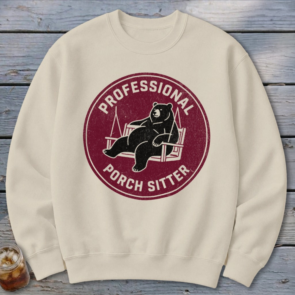 Beige crewneck sweatshirt with a maroon circular graphic featuring a bear relaxing on a porch swing and the text "PROFESSIONAL PORCH SITTER".