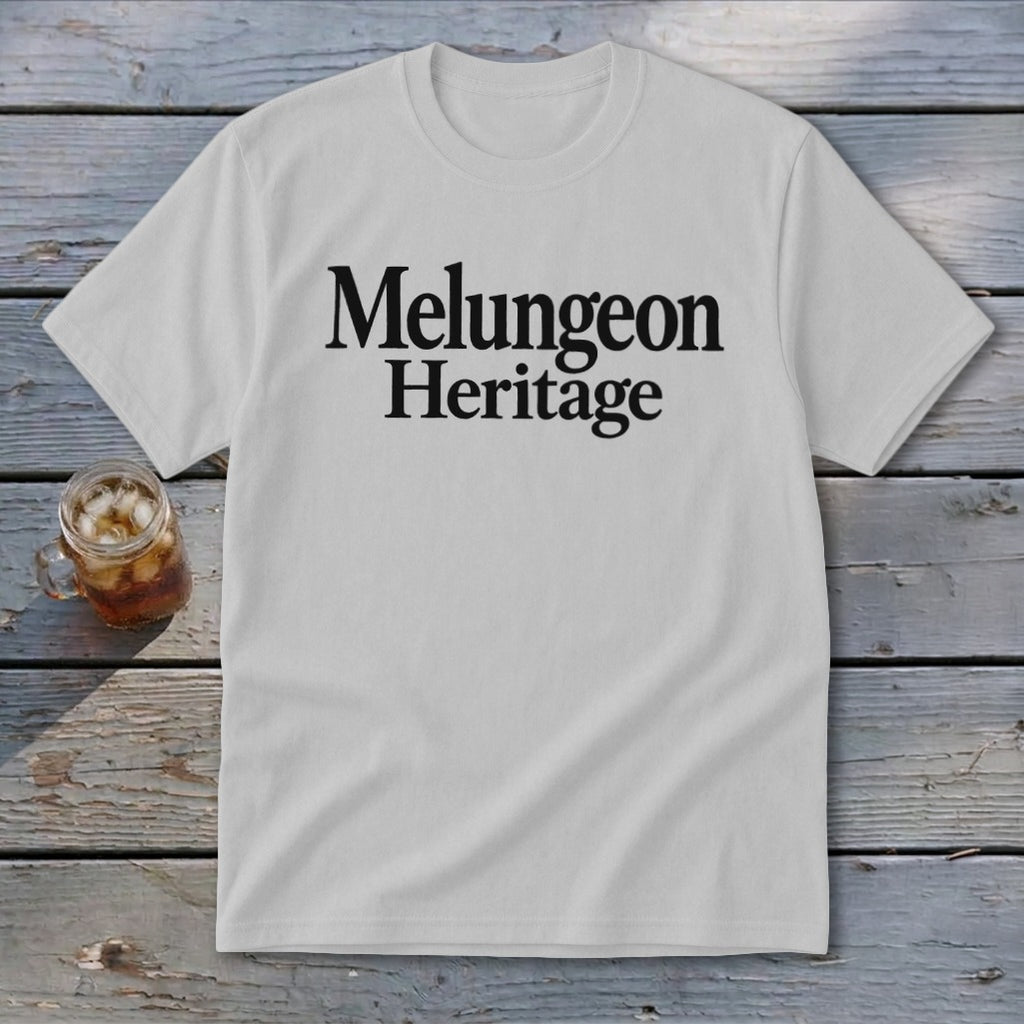 A light gray crewneck t-shirt featuring the black text Melungeon Heritage laid flat on a rustic wooden surface next to a glass of iced tea.