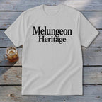 A light gray crewneck t-shirt featuring the black text Melungeon Heritage laid flat on a rustic wooden surface next to a glass of iced tea.