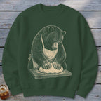 Bear wearing an apron kneading dough on a wooden cutting board.