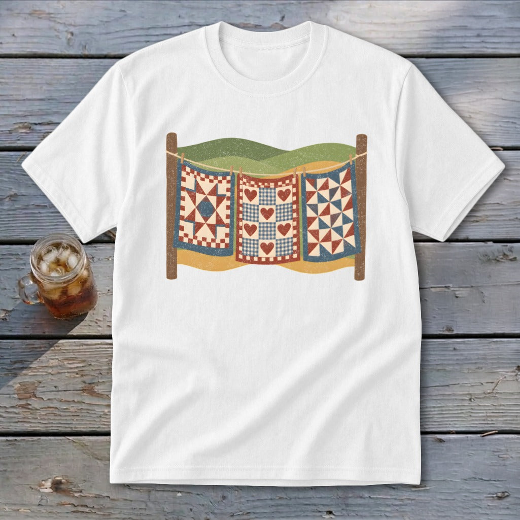 A white t-shirt featuring a graphic of three colorful quilts hanging on a clothesline against a landscape.