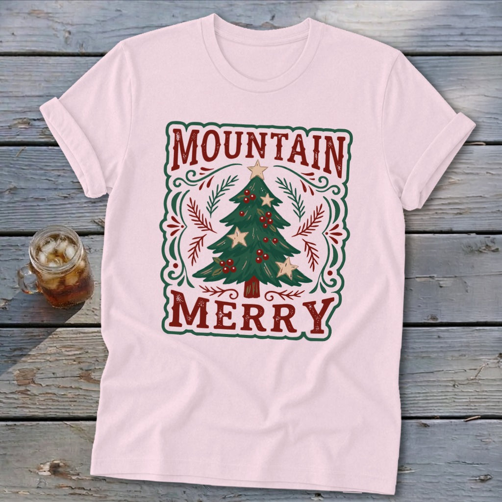 A light pink t-shirt with a vintage Christmas tree design and the text Mountain Merry on a rustic wooden background.