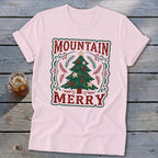 A light pink t-shirt with a vintage Christmas tree design and the text Mountain Merry on a rustic wooden background.
