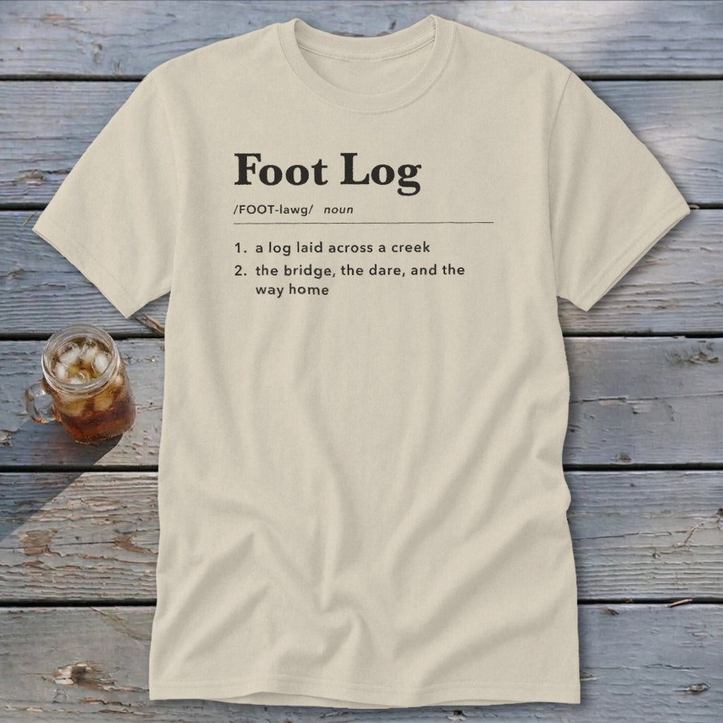 A cream-colored crewneck t-shirt featuring dictionary-style text defining Foot Log lies flat on a weathered wooden surface next to an iced beverage.