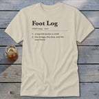 A cream-colored crewneck t-shirt featuring dictionary-style text defining Foot Log lies flat on a weathered wooden surface next to an iced beverage.