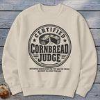 Beige crewneck sweatshirt featuring a black graphic with a round badge, a round cornbread loaf, and the text "Certified Cornbread Judge".