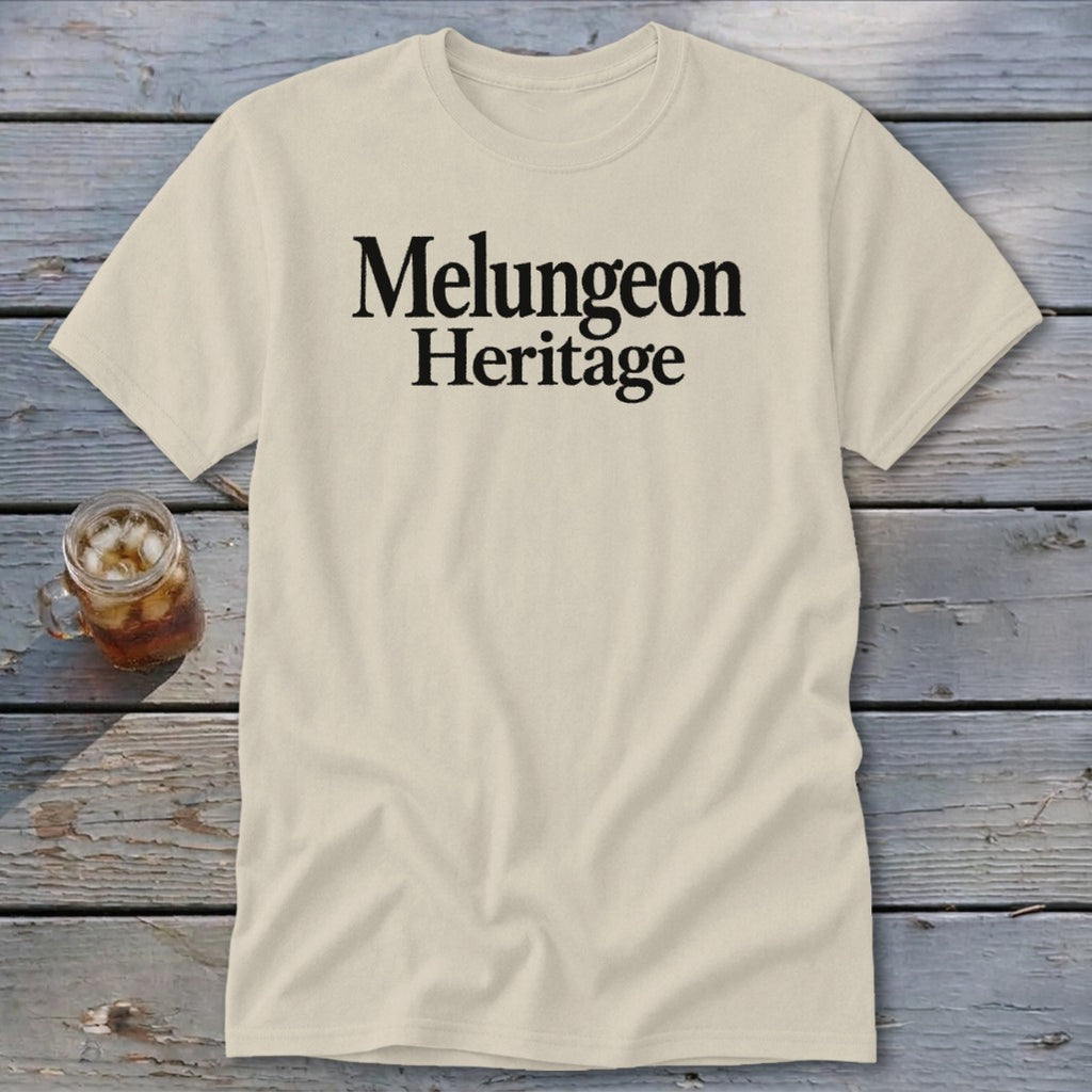 A cream-colored t-shirt with the black text "Melungeon Heritage" centered on the chest, laid flat on a rustic wooden surface next to a glass of iced tea.