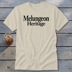 A cream-colored t-shirt with the black text "Melungeon Heritage" centered on the chest, laid flat on a rustic wooden surface next to a glass of iced tea.