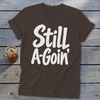 A brown short-sleeve t-shirt with rolled cuffs and white text that reads Still A-Goin'.