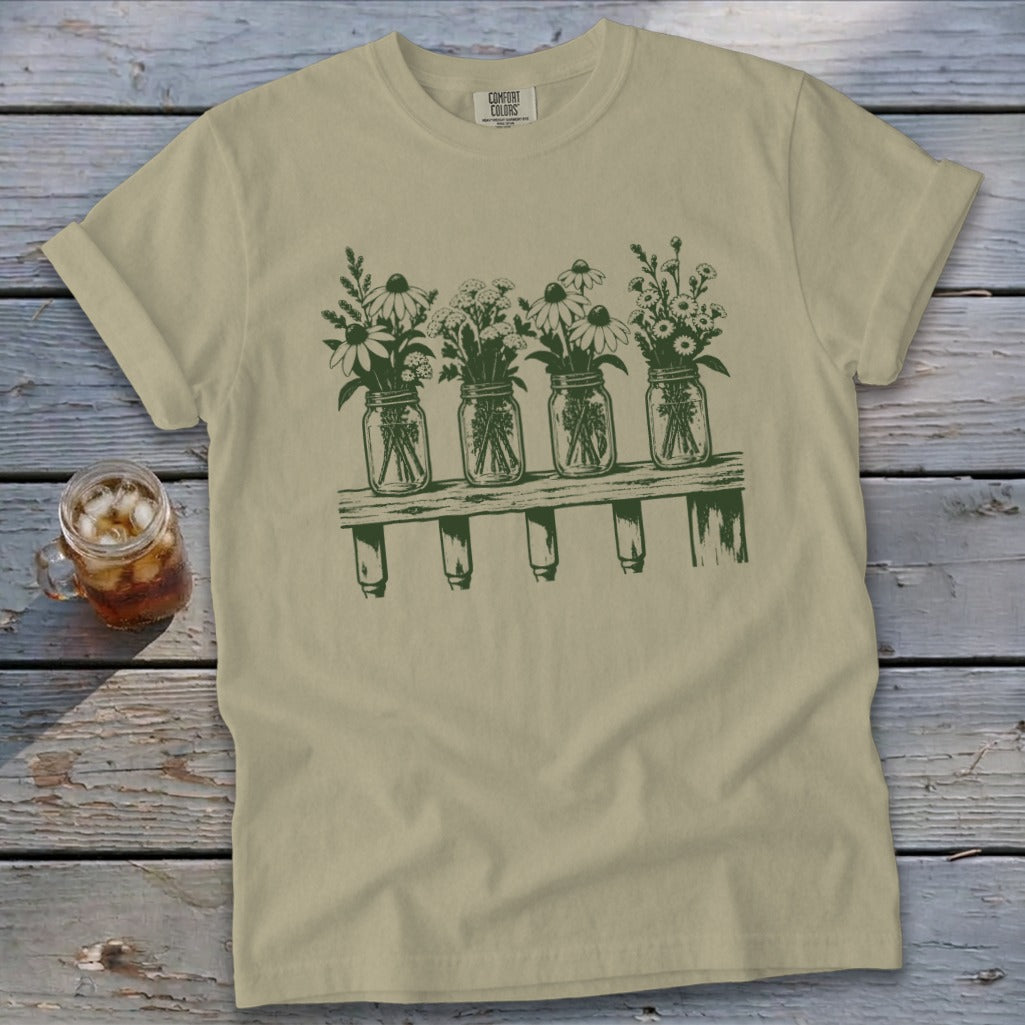A light olive green t-shirt with a green graphic of four mason jars filled with wildflowers on a shelf.