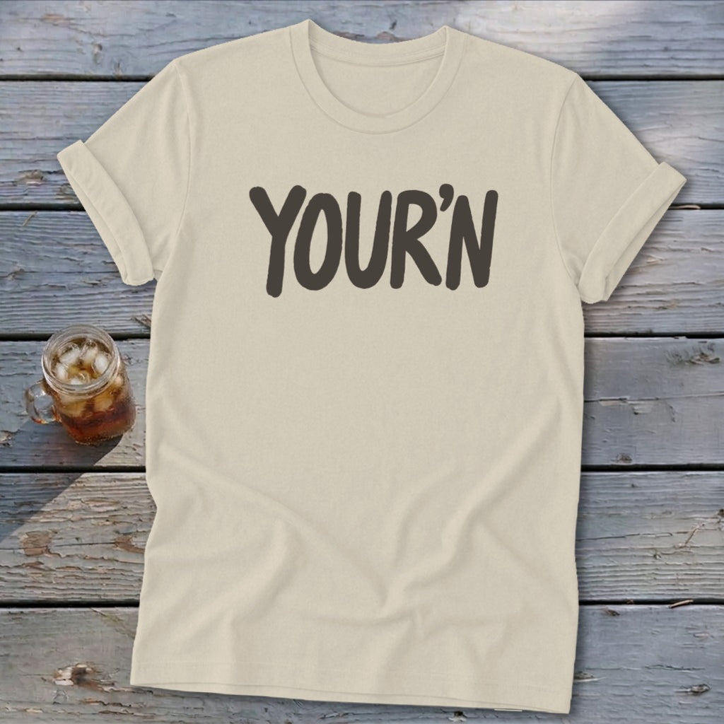 Cream t-shirt with "YOUR'N" text in dark gray, placed on a wooden surface.