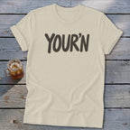 Cream t-shirt with "YOUR'N" text in dark gray, placed on a wooden surface.