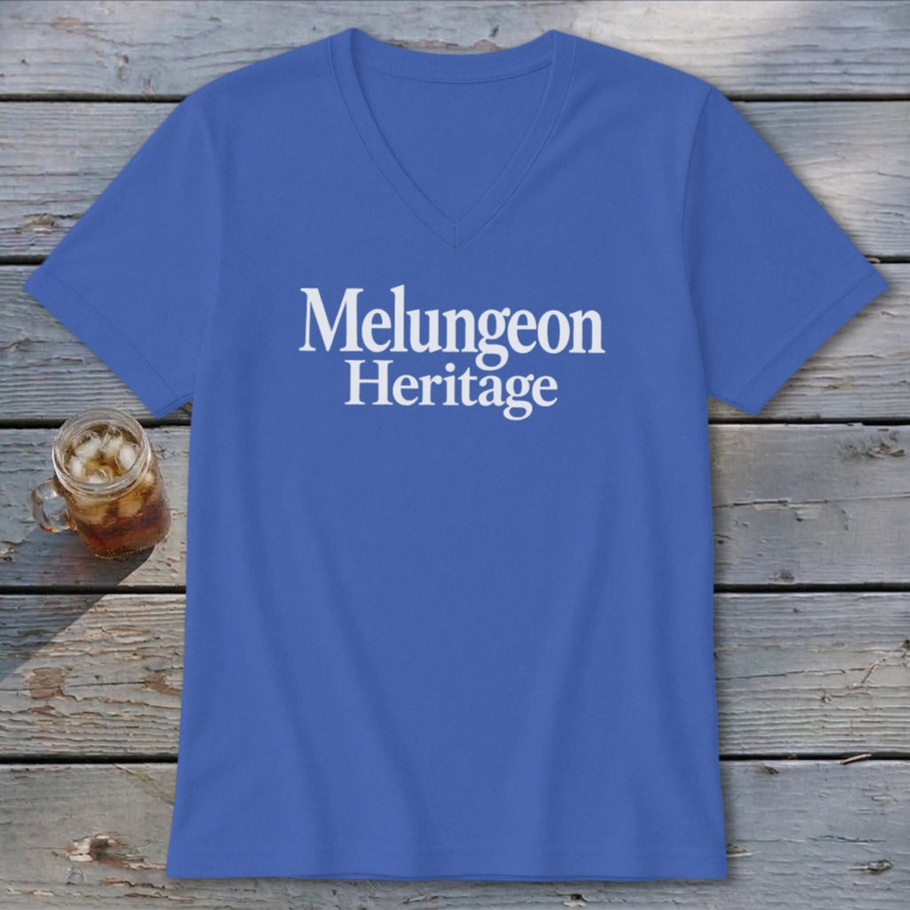 Blue V-neck t-shirt with white text reading Melungeon Heritage centered on the chest, laid flat on a rustic wooden table next to a glass of iced tea.