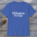 Blue V-neck t-shirt with white text reading Melungeon Heritage centered on the chest, laid flat on a rustic wooden table next to a glass of iced tea.