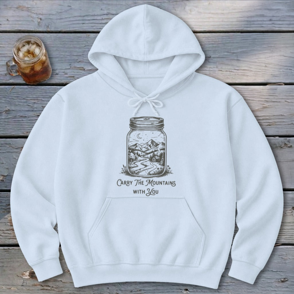 A white hooded sweatshirt featuring a line-art graphic of a mountain scene inside a mason jar and the text "Carry The Mountains With You."