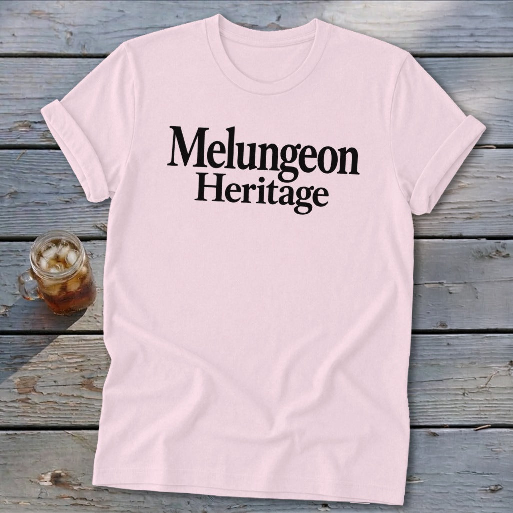 A light pink t-shirt with rolled sleeves displays the black text Melungeon Heritage across the chest, laid flat on a rustic wooden surface next to a glass of iced tea.