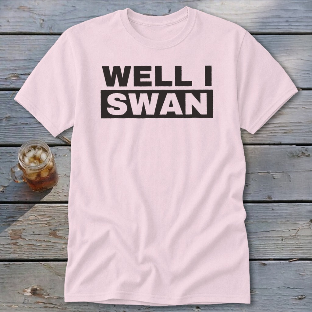 A light pink t-shirt with black block lettering reading WELL I SWAN lies on a weathered wooden surface next to a glass of iced tea.