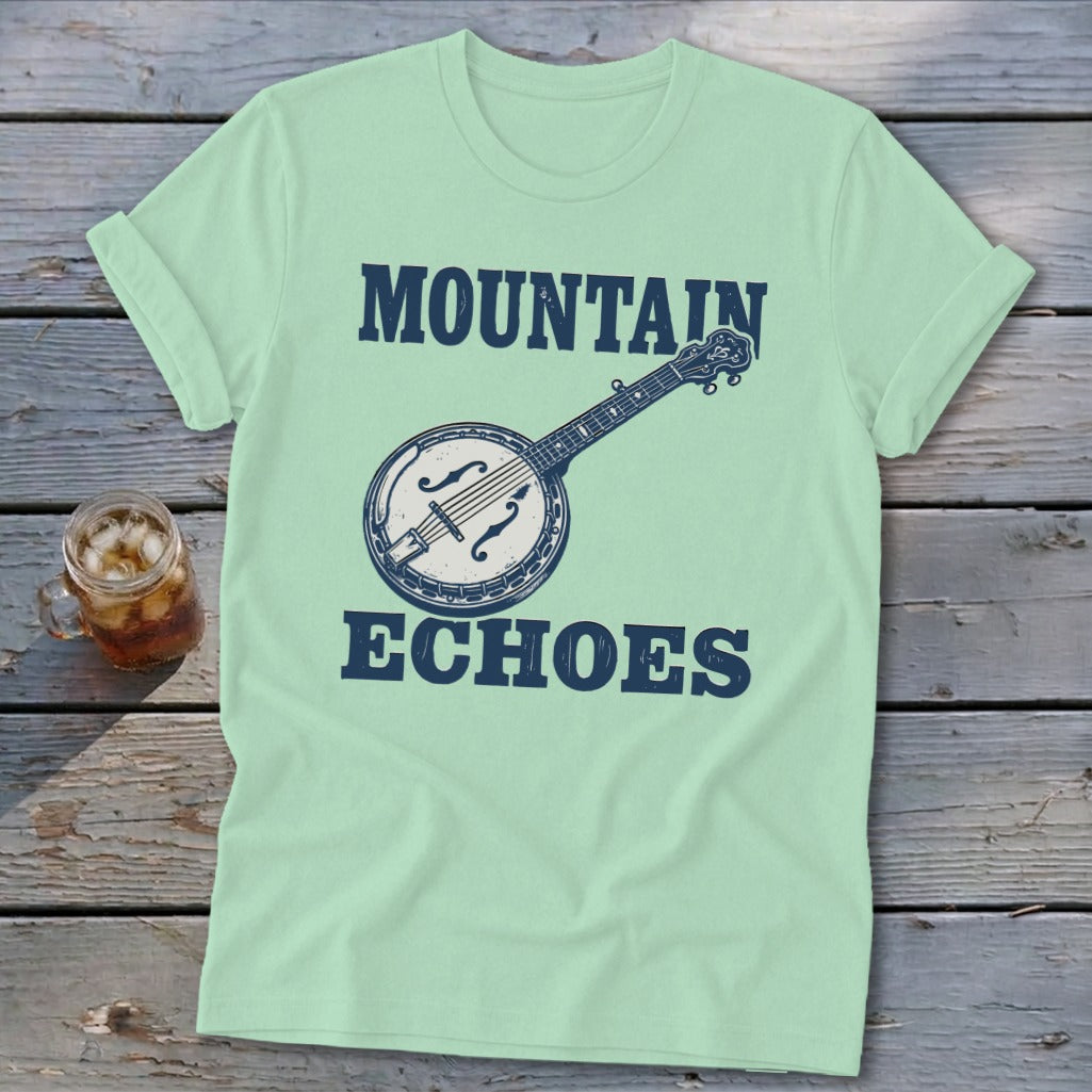 Light green t-shirt with a vintage banjo graphic and text reading Mountain Echoes, laid on wood.