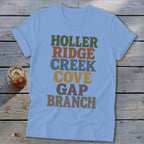 Light blue t-shirt featuring stacked multicolored text reading Holler Ridge Creek Cove Gap Branch, laid on wooden planks.