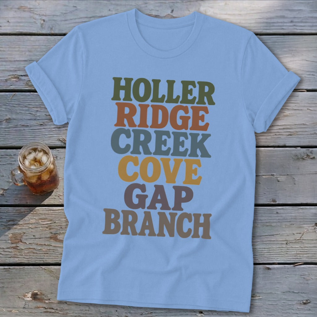 Light blue t-shirt featuring stacked multicolored text reading Holler Ridge Creek Cove Gap Branch, laid on wooden planks.