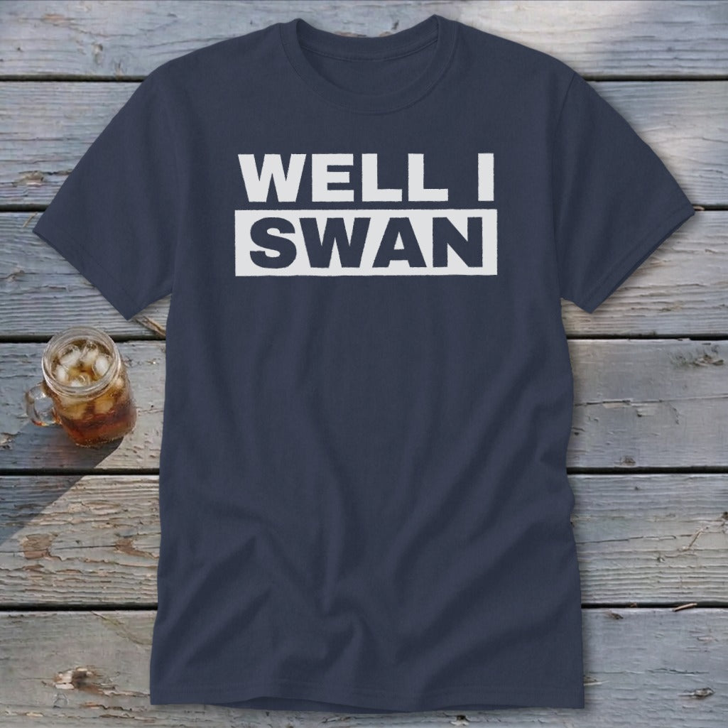 A dark blue t-shirt printed with the white text WELL I SWAN lying on a weathered wooden surface next to a glass of iced tea.