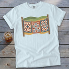 Light blue t-shirt featuring a graphic of three vintage quilts hanging on a clothesline outdoors.