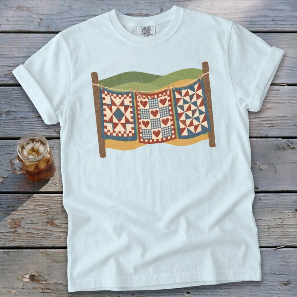 Light blue t-shirt featuring a graphic of three vintage quilts hanging on a clothesline outdoors.