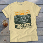 Light yellow t-shirt with a graphic of mountains and sunrise, text "RIDGELINE AT DAYBREAK".