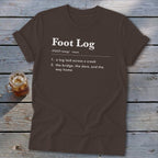 Brown t-shirt featuring dictionary-style text defining Foot Log laid across a wooden surface.