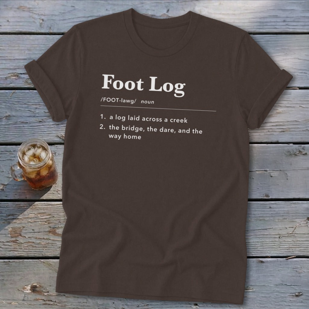 Brown t-shirt featuring dictionary-style text defining Foot Log laid across a wooden surface.