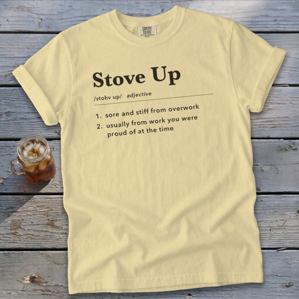 A light yellow t-shirt laid flat on a wooden surface, featuring black text defining the term stove up.