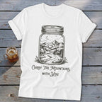 A white t-shirt featuring a mason jar graphic containing a mountain landscape and the text Carry The Mountains With You.