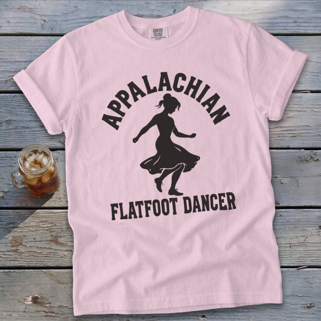 Light pink t-shirt with black text reading Appalachian Flatfoot Dancer surrounding a dancer's silhouette, laid flat on a rustic wooden surface.