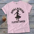 Light pink t-shirt with black text reading Appalachian Flatfoot Dancer surrounding a dancer's silhouette, laid flat on a rustic wooden surface.