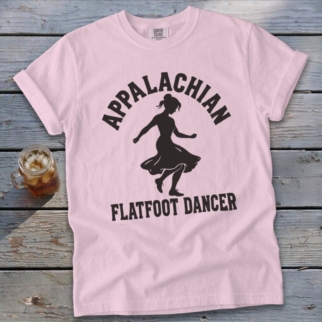 Light pink t-shirt with black text reading Appalachian Flatfoot Dancer surrounding a dancer's silhouette, laid flat on a rustic wooden surface.