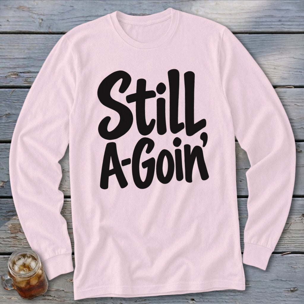 Light pink long-sleeved shirt with black lettering reading Still A-Goin' on a wooden background.