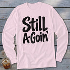 Light pink long-sleeved shirt with black lettering reading Still A-Goin' on a wooden background.