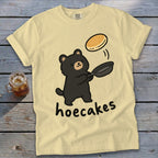 A yellow t-shirt featuring a cartoon bear flipping a pancake from a skillet, with the text hoecakes.