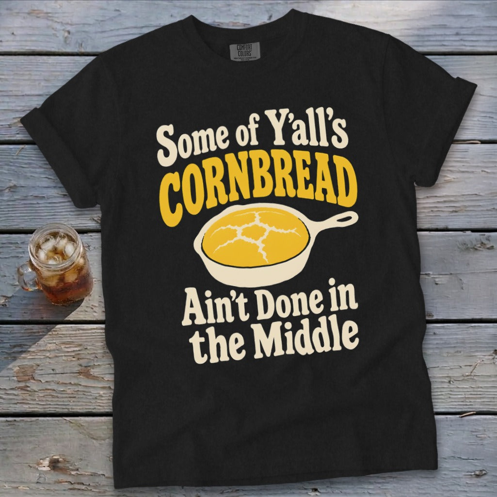 A black t-shirt with the text "Some of Y'all's CORNBREAD Ain't Done in the Middle" surrounding a skillet illustration.