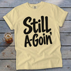 A yellow t-shirt with black cursive text reading Still A-Goin' on a wooden surface.