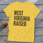 Yellow t-shirt with "WEST VIRGINIA RAISED" text, on a wooden surface.