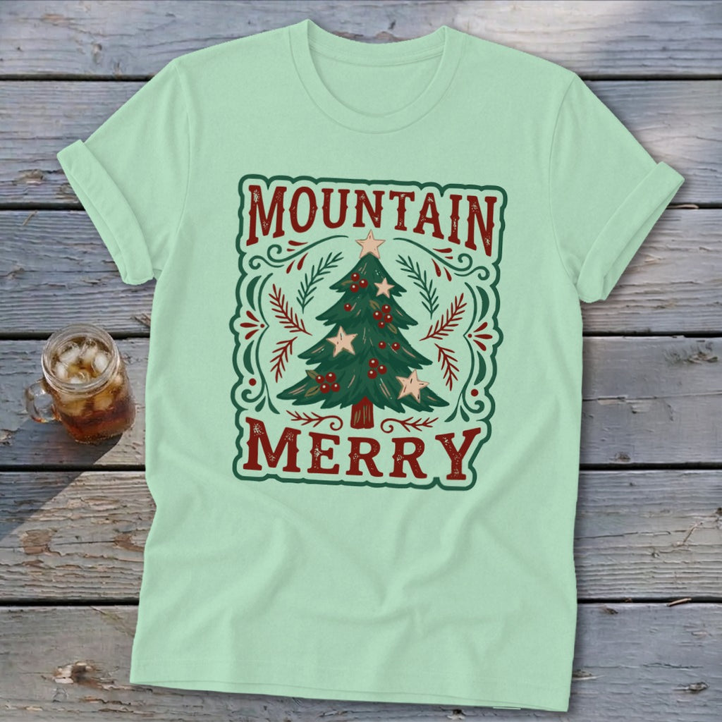 Light green t-shirt featuring a Christmas graphic with the text Mountain Merry on a wooden surface.