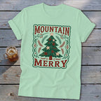 Light green t-shirt featuring a Christmas graphic with the text Mountain Merry on a wooden surface.