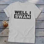 A light gray t-shirt featuring the black text Well I Swan across the chest, laid flat on a rustic wooden surface next to a glass of iced tea.