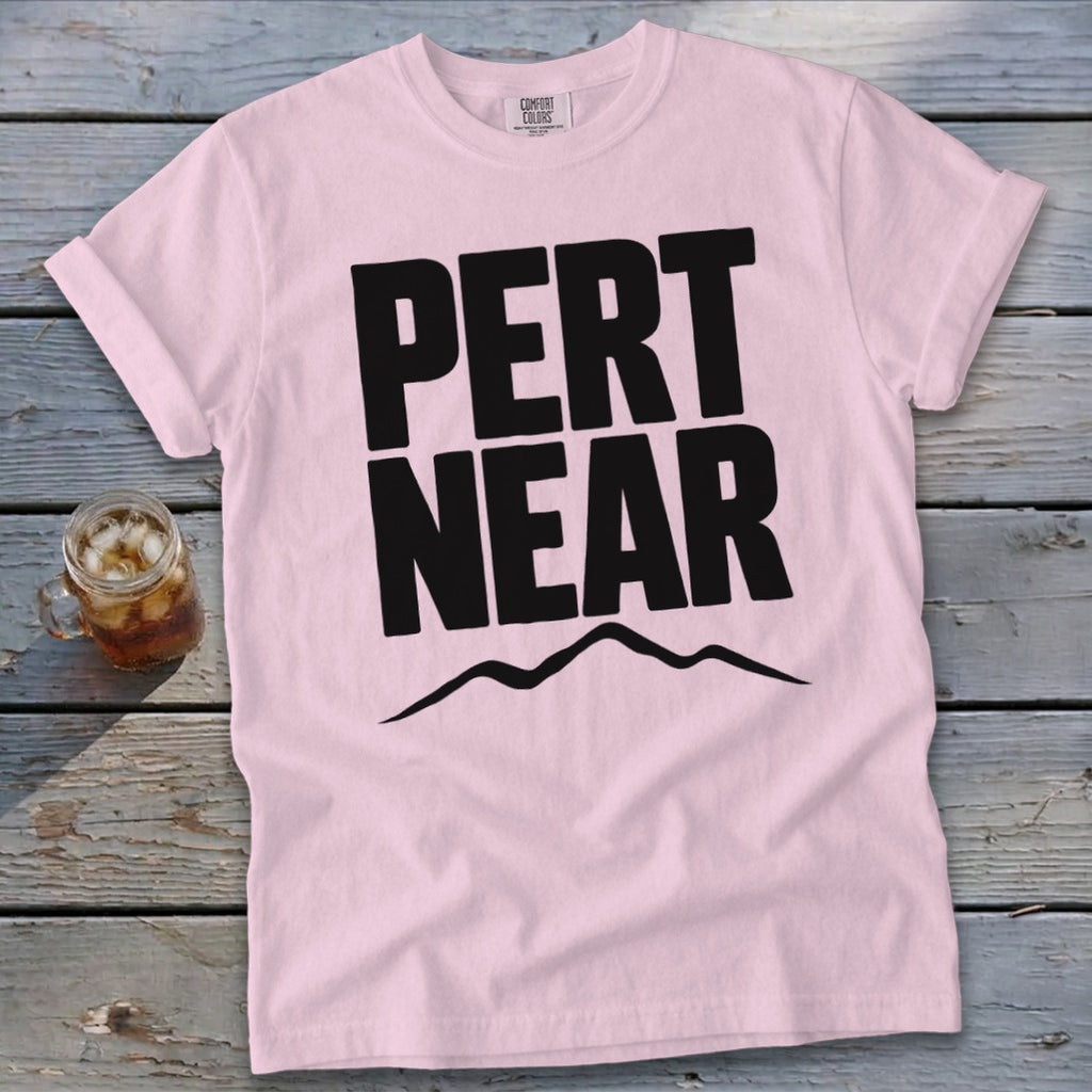 Light pink t-shirt featuring the black text PERT NEAR above a mountain range graphic, resting on a weathered wooden surface next to an iced beverage.