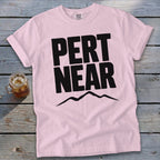 Light pink t-shirt featuring the black text PERT NEAR above a mountain range graphic, resting on a weathered wooden surface next to an iced beverage.